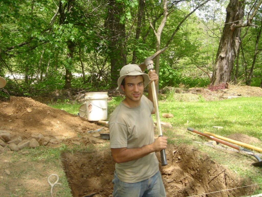 Bill Burns in the field as an archaeologist, standing with a pick beside an excavation unit with string grid lines, a sifting screen, and a spoil pile.